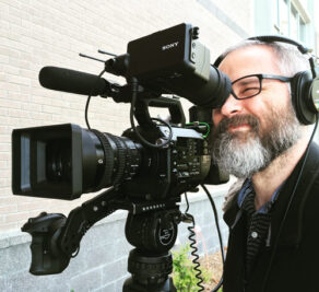 A bespectacled Greg Winters of Kino Mountain Productions looking into the viewfinder of his video camera