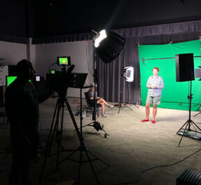 A woman standing in front of a green screen with video cameras and production equipment in front of her.